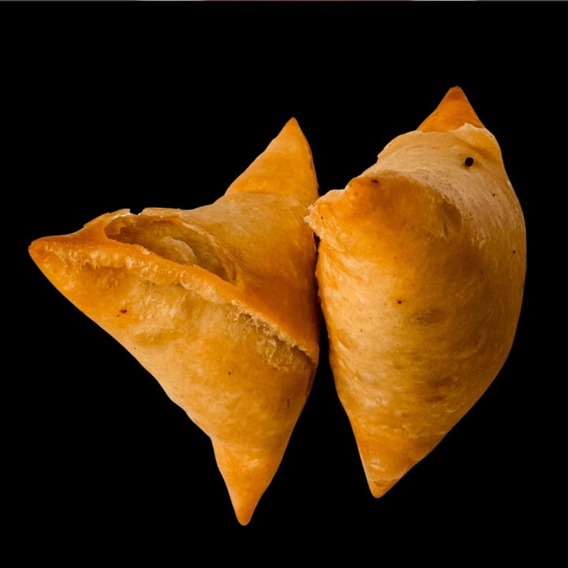 Two golden-brown Dhaba Kitchen samosas, one slightly open, against a dark background.